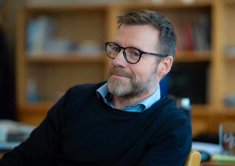 Foto av Åsmund Sandvik, kommunedirektør i Øyer kommune. Han har kort mørkt hår, skjegg og briller, og har på seg en mørk genser med en blå skjorte under. Han smiler.