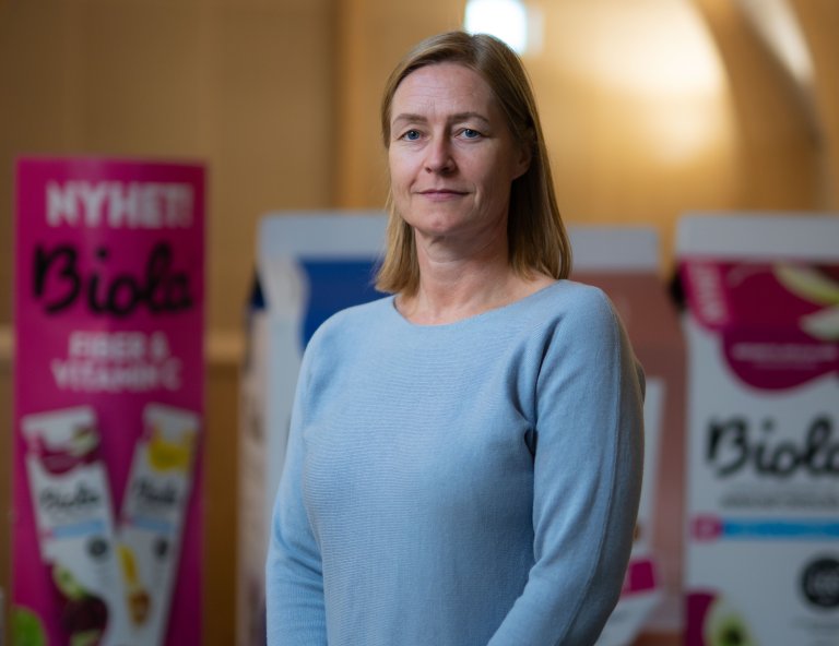 Portrettfoto av Lene Grøndal. Hun står foran plansjer med reklame for ulike TINE-produkter. Hun har på seg en blå genser.