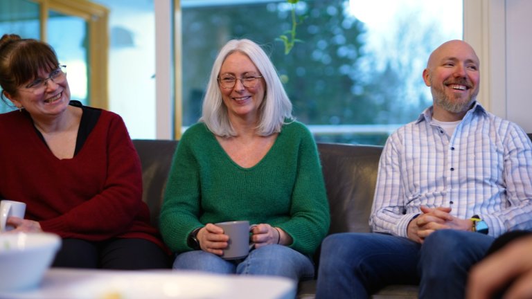 Foto av to kvinner og en mann som sitter sammen i en sofa. De smiler og ler.